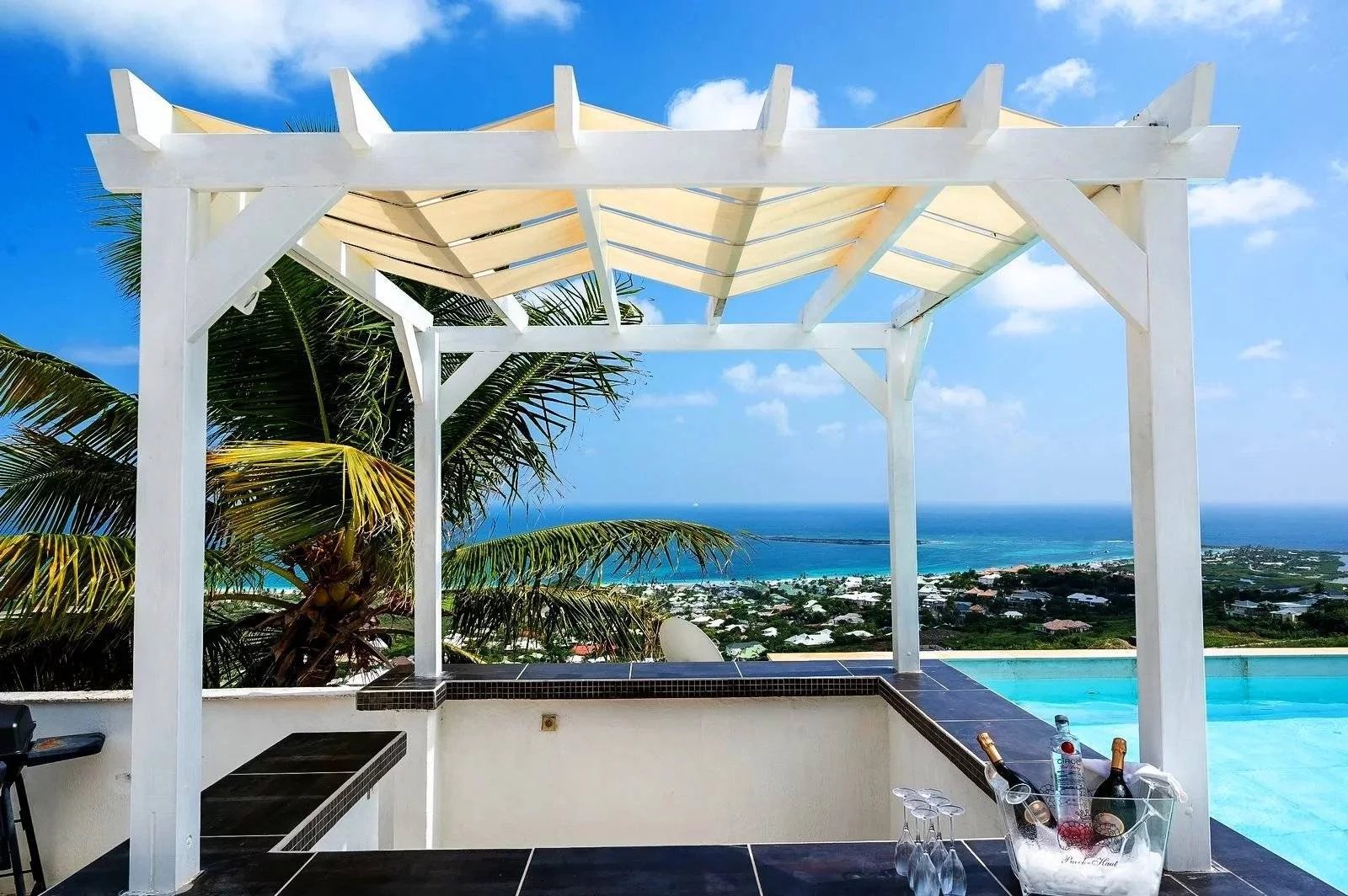 White wooden pergola over a dark-tile bar with a bottle bucket, overlooking the blue ocean and palm trees.