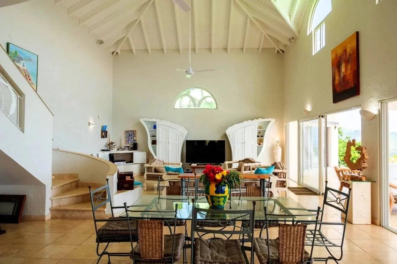 Bright open living and dining area with a glass dining table, wicker chairs, a colorful vase, and a TV between white curved cabinets.