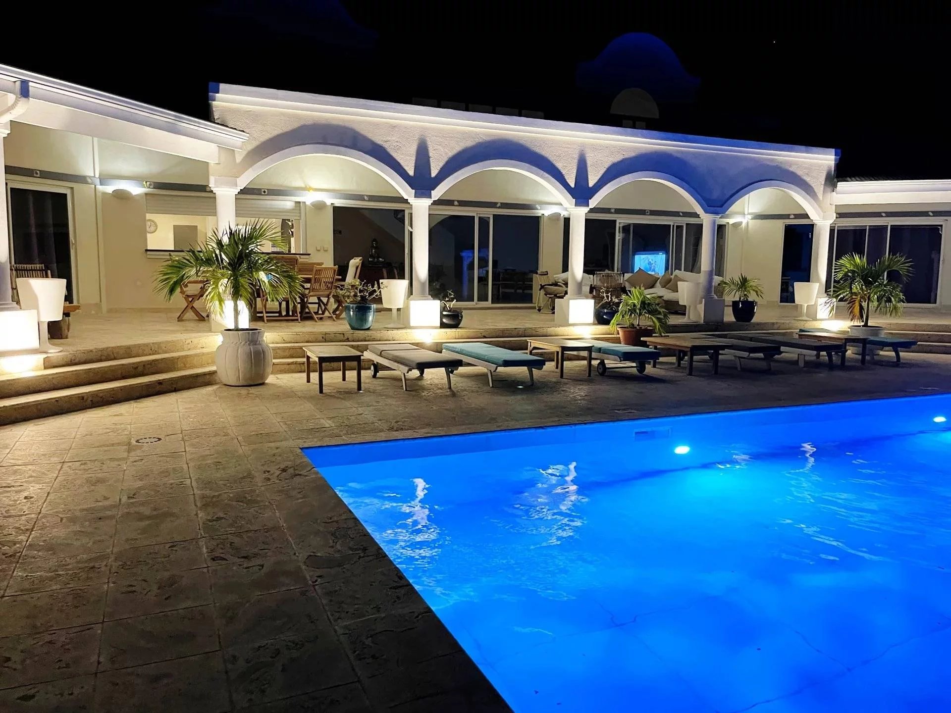 Nighttime poolside at a villa with blue-lit rectangular pool, white arches, and lounge chairs along a tiled deck.
