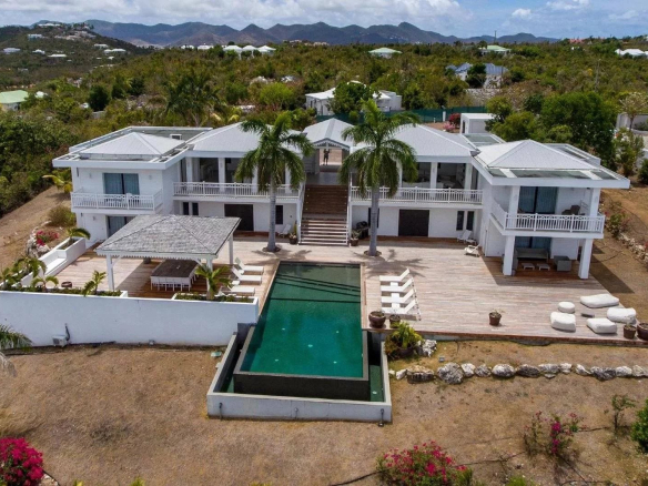 Aerial view of a large white two-story villa with palm trees, broad wooden deck, and a central rectangular pool.