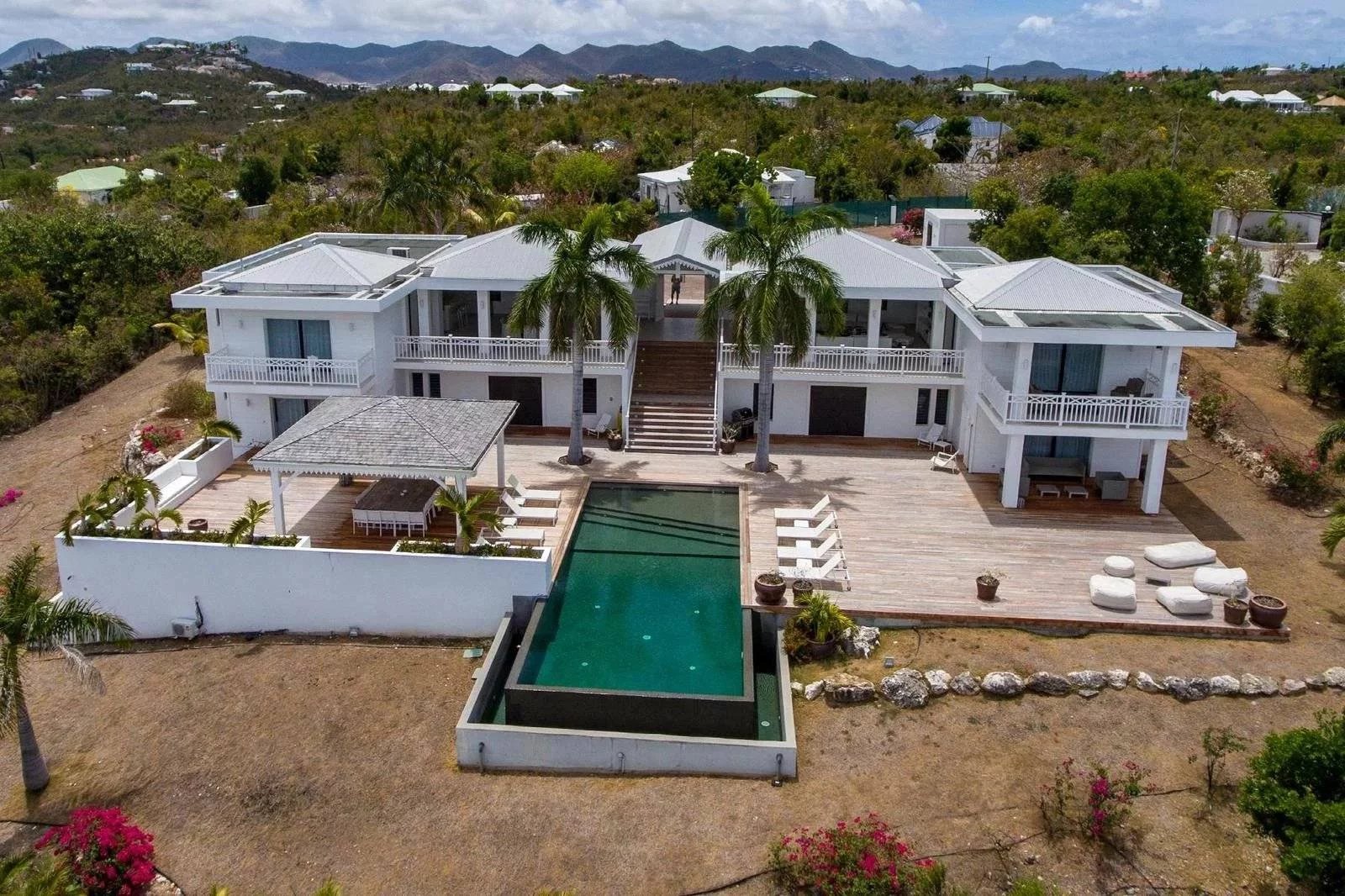 002e8f322f9d6138271604c29d921300_f9b5d9e344_1600-original Aerial view of a large white two-story villa with palm trees, broad wooden deck, and a central rectangular pool.