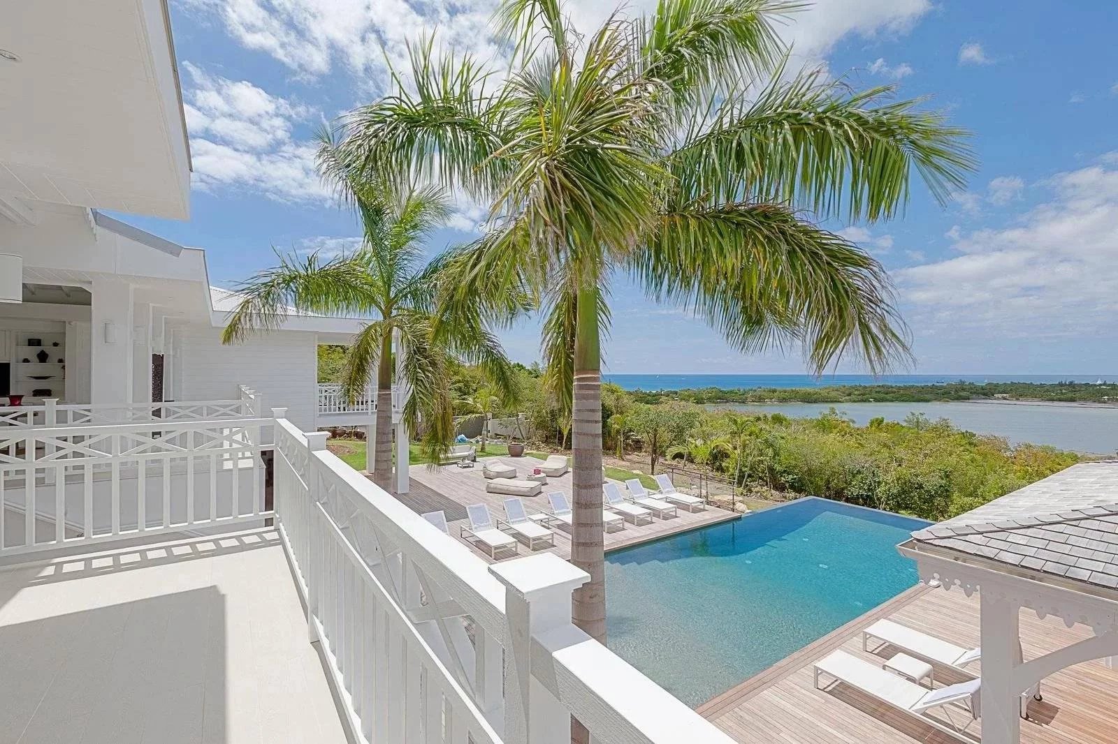 2418b54791f161dd462812acb9677e83_138da6e832_1600-original White coastal villa with a spacious balcony, palm trees, and a turquoise pool overlooking the ocean under a bright blue sky.