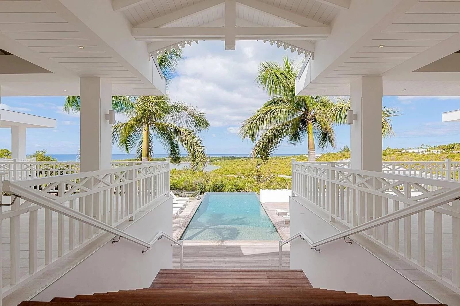 3cde7873932e18d2a0f81aa160037d24_e58b452af2_1600-original White beachfront resort veranda with wooden stairs to a rectangular pool; palm trees and the ocean beyond.