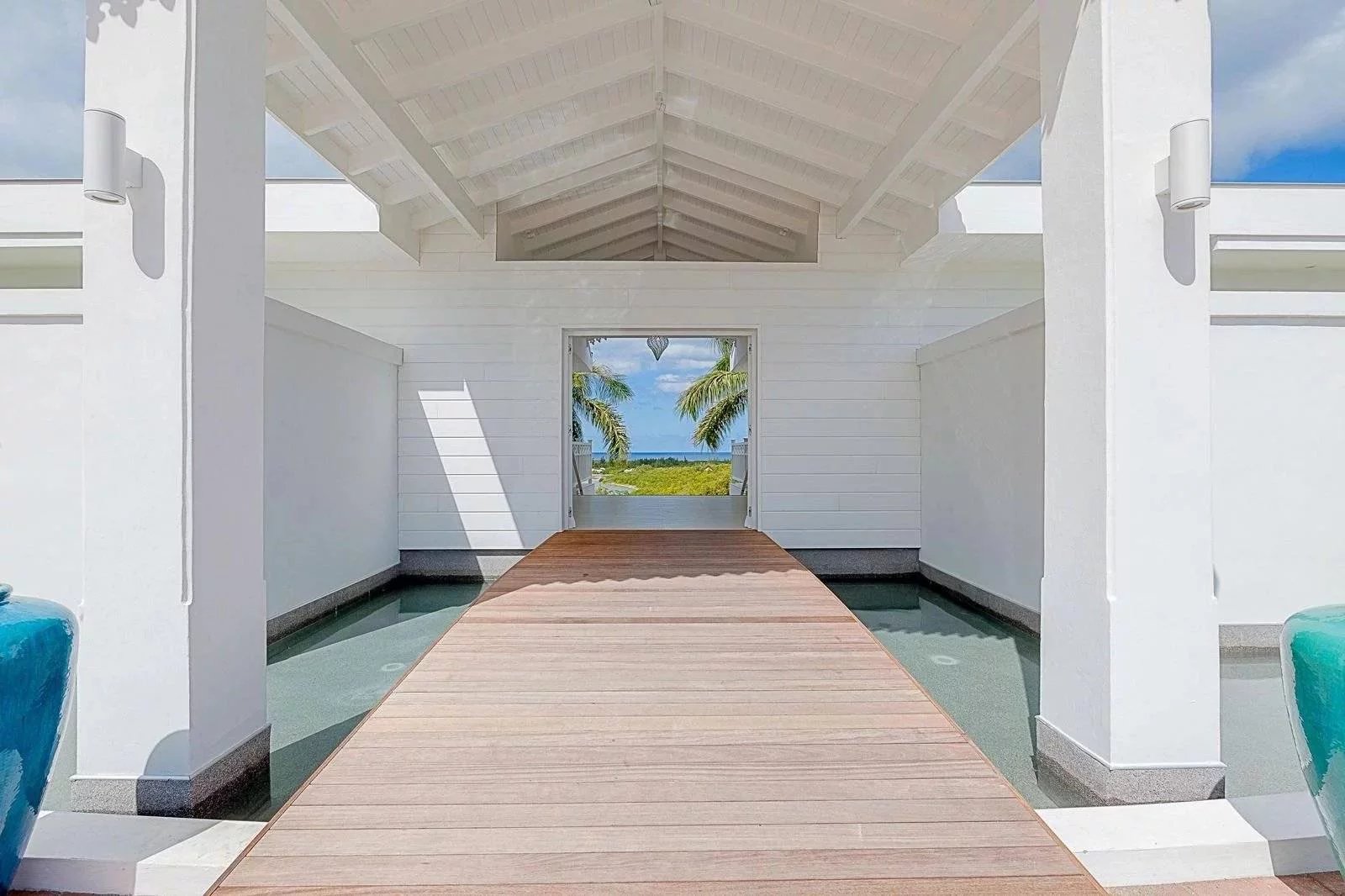 439cd258c08314ce72aca6eb2a73a3d6_6bba76d4da_1600-original White seaside pavilion with a wooden boardwalk leading to a beach through an open doorway, palms in view beyond the entrance.