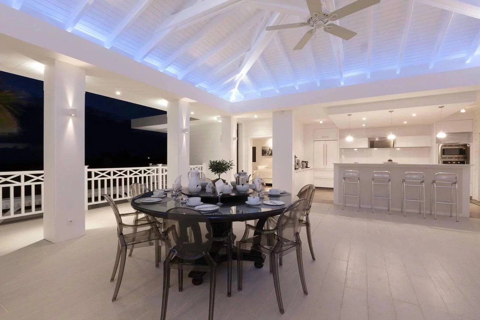 450fa049ef2fea76ee57b7f4eaabb98b_7c68aa43fd_1600-original White open-air dining area at night with a round glass table set for eight and a white kitchen island in the background.