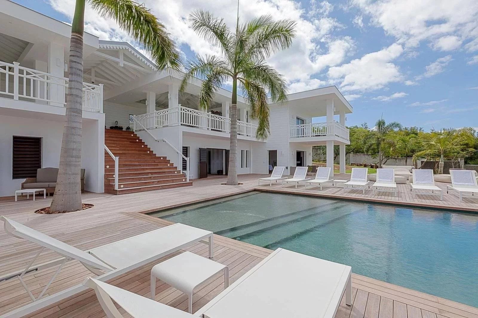 680f6a8fa75a6b6965cf32b4704f3aa5_e6f4e86693_1600-original White modern villa with a rectangular pool, wooden deck, and palm trees under a blue sky with lounge chairs nearby.