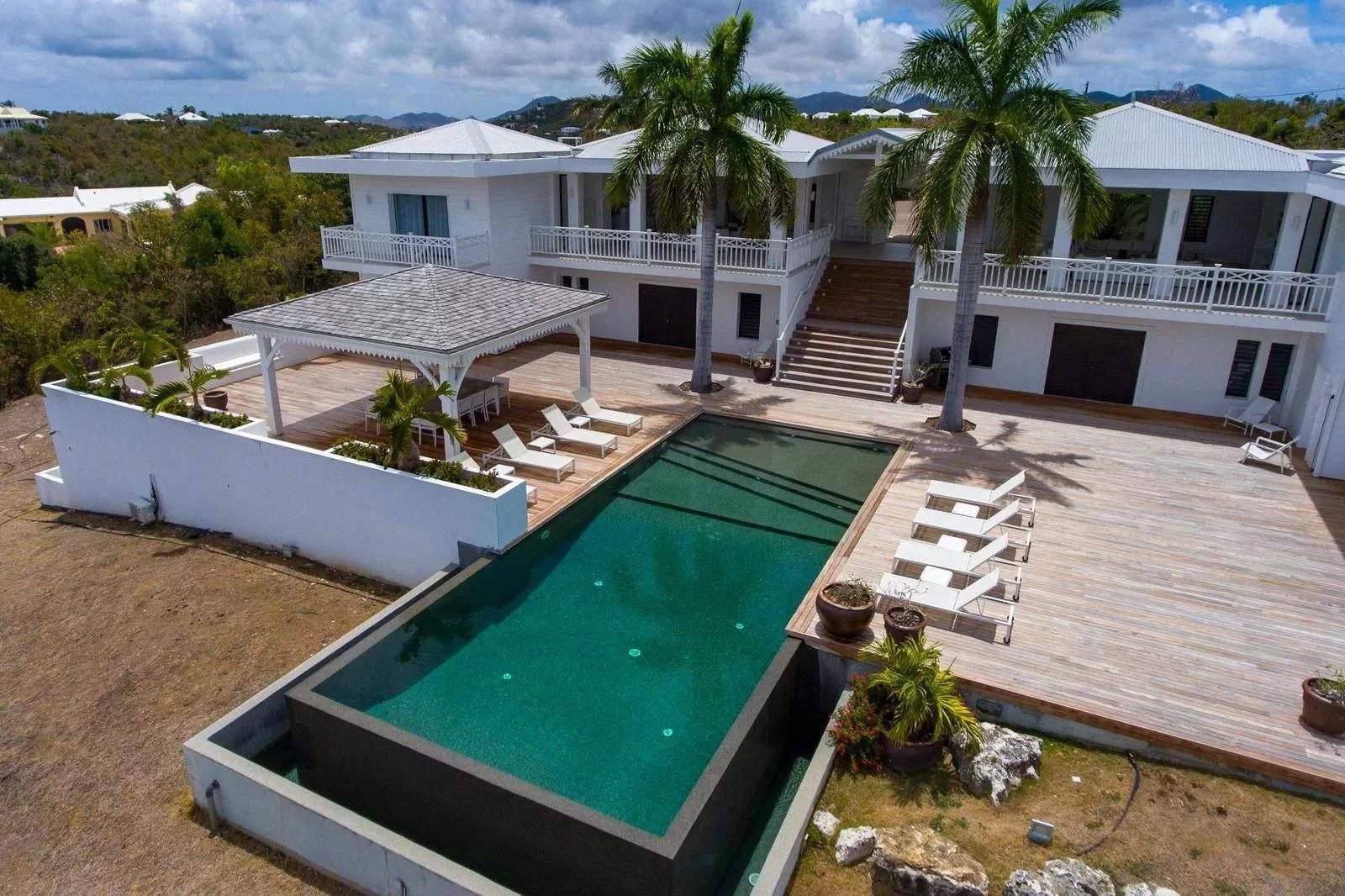 89c642458c3028c4f3de64cc6c4118a5_bee82670eb_1600-original Aerial view of a white two‑story villa with a rectangular turquoise pool, wooden deck, and palm trees with lounge chairs nearby.