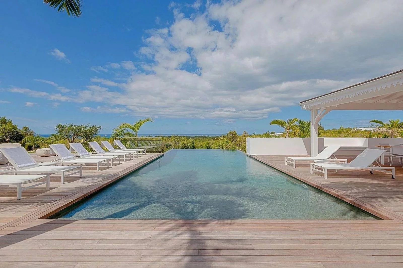 92be7a51473e44b9bdb18c51bae5bb20_dc16eba5d6_1600-original Rectangular outdoor pool on a wooden deck with white lounge chairs, a shaded seating area to the right, and a tropical landscape under a blue sky.