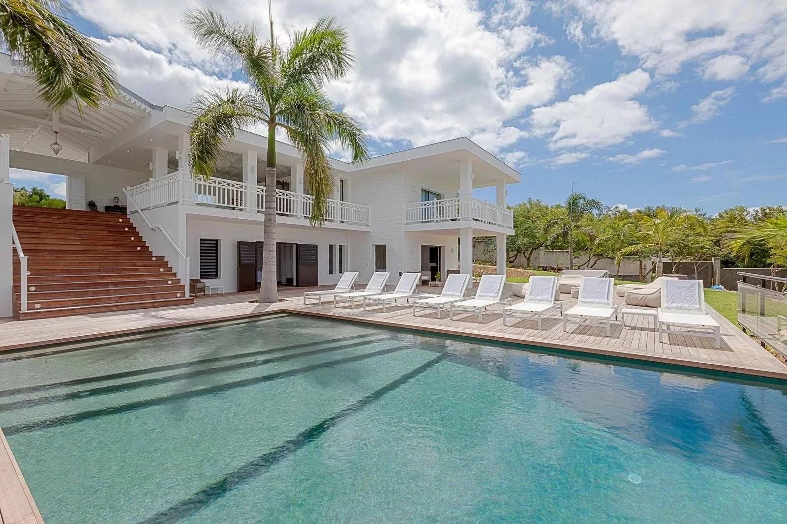 bbe3cc65ed766344853a005c2612bc43_e9839b09cc_1600-original White two-story villa with a rectangular pool, wooden deck, and row of white lounge chairs under palm trees on a sunny day.