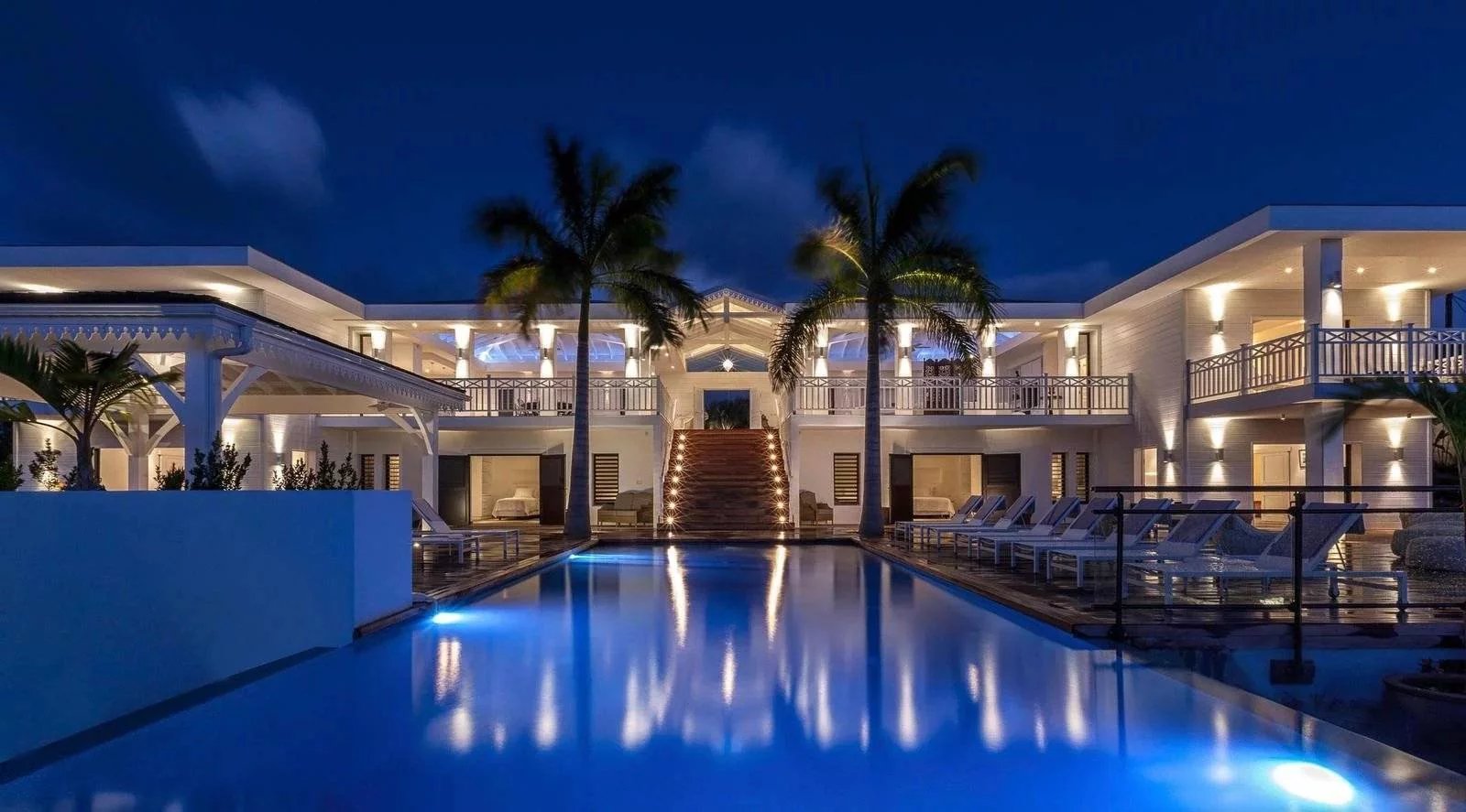 d20382ef86c34b1f16c221ae064bb0b5_9f39fb3629_1600-original Nighttime view of a luxury resort with a central illuminated staircase, palm trees, and a blue-lit pool reflecting the buildings.