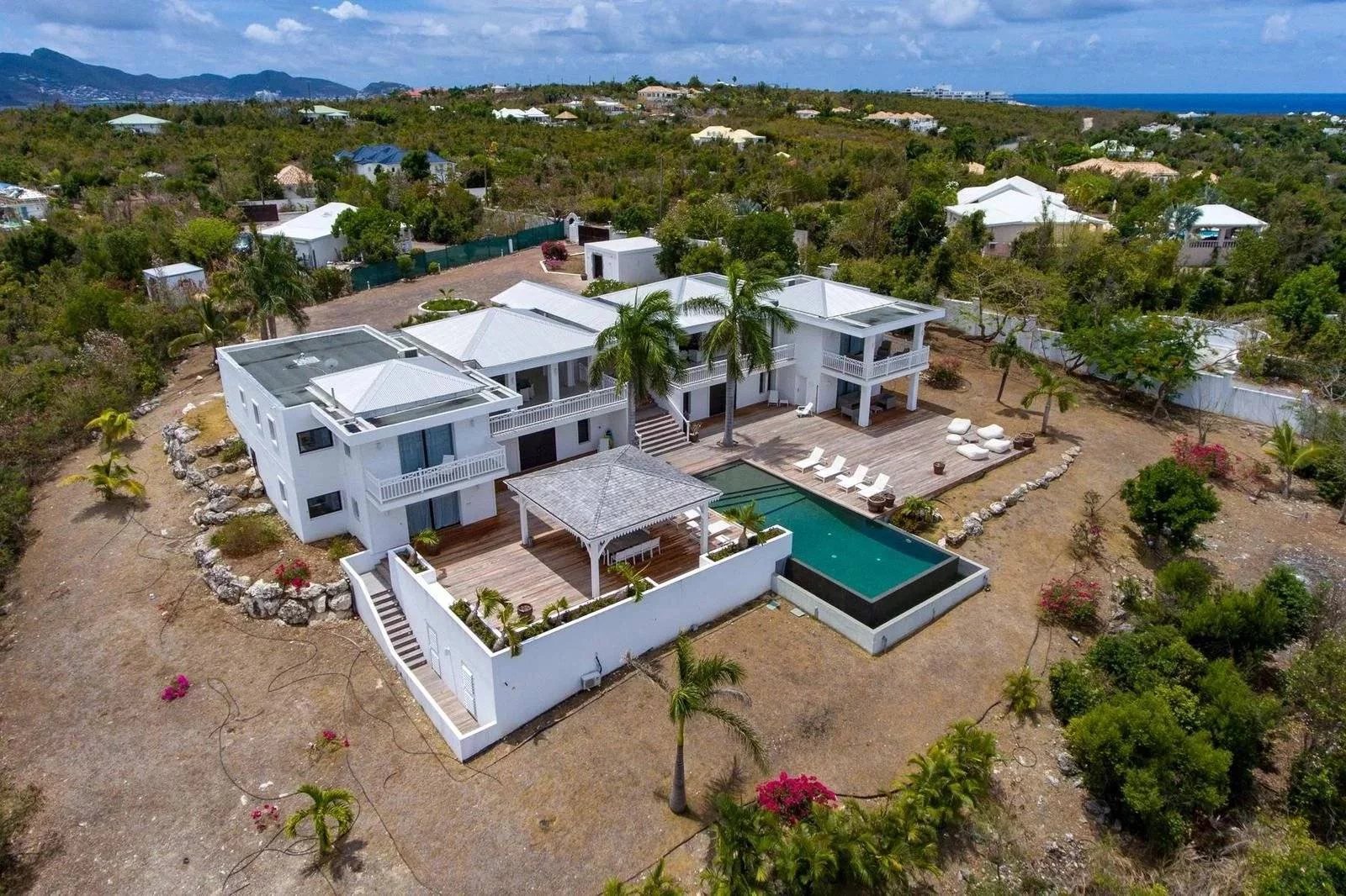 d37ed54e1edfbe17aefc100938b10483_95345e564d_1600-original Aerial view of a white two-story villa with a rectangular pool and wooden deck in a tropical yard.