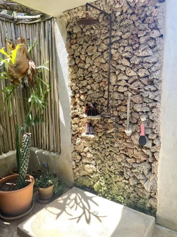 Outdoor shower area with a stone wall, overhead rain showerhead, and a small shelf holding bottles; garden pots and a cactus nearby.