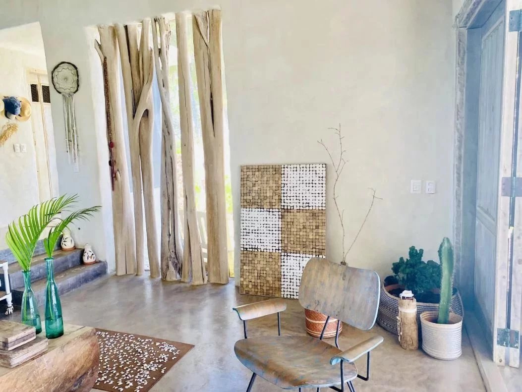 Sunlit boho living room corner with a driftwood screen, a wooden chair, and potted plants plus a mosaic tile panel against the wall.