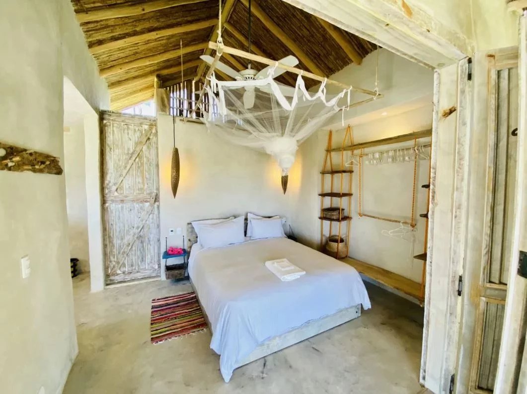 Rustic bedroom with a white bed, red striped rug, and woven wooden shelves against a plastered wall under a thatched ceiling. A web-like canopy hangs above the bed.