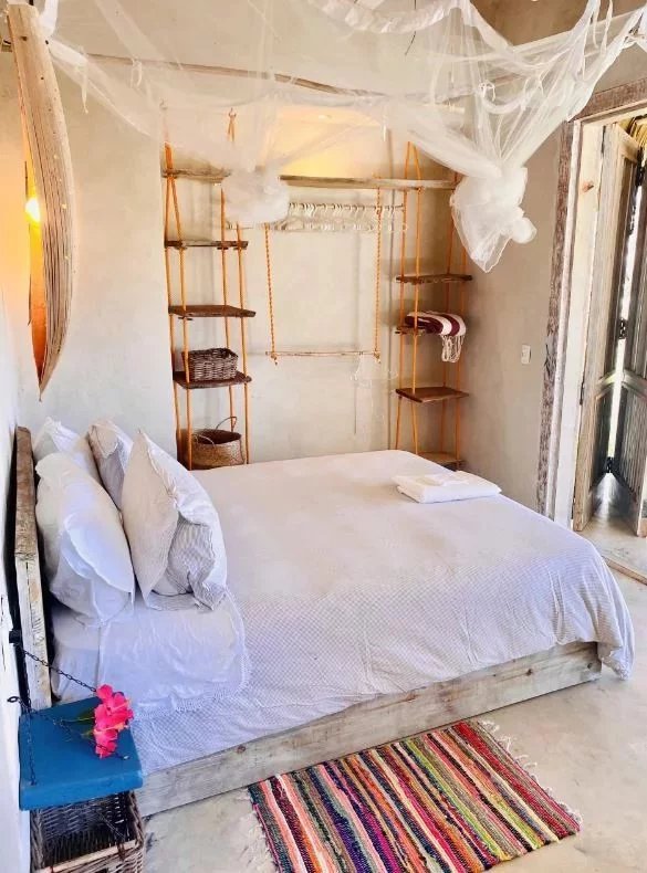 Rustic bedroom with a wooden bed, white bedding, and a net canopy; open shelves and wicker baskets along the back wall.