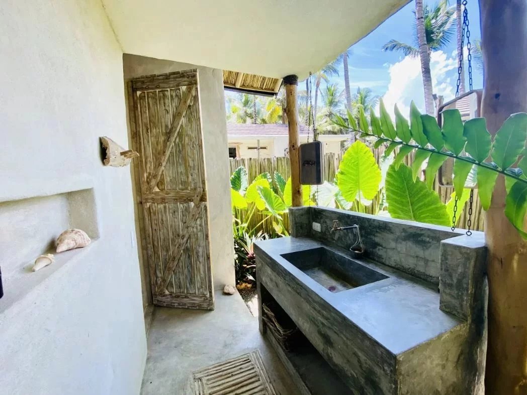 Outdoor concrete double sink with stone countertop under a covered patio, weathered wooden door to the left, tropical plants in the background.