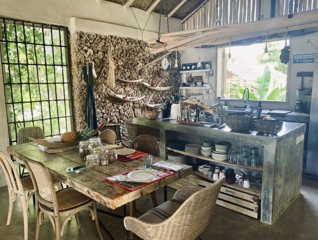Rustic indoor–outdoor dining and kitchen area with a weathered wooden table, wicker chairs, a stone wall, and a concrete island with dishes and glassware.