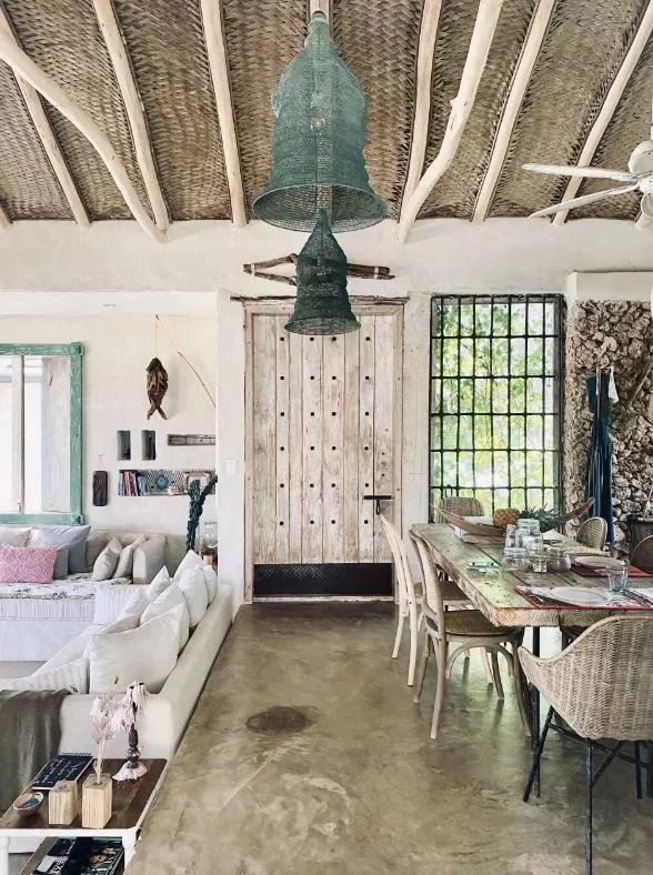 Rustic open-plan living and dining area with large green bell-shaped pendant lights, white sectional sofa, and a weathered wooden door in the center.