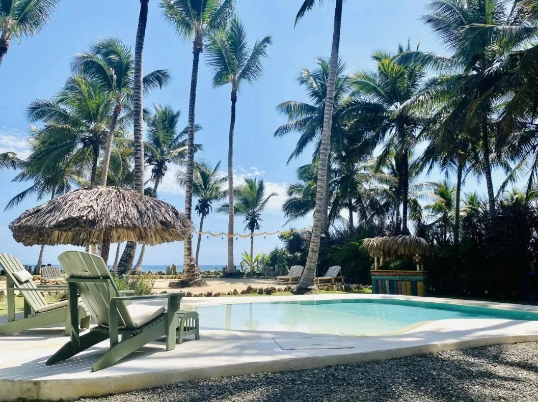 Poolside scene with thatched umbrellas, green lounge chairs, and tall palm trees under a blue sky at a tropical resort.