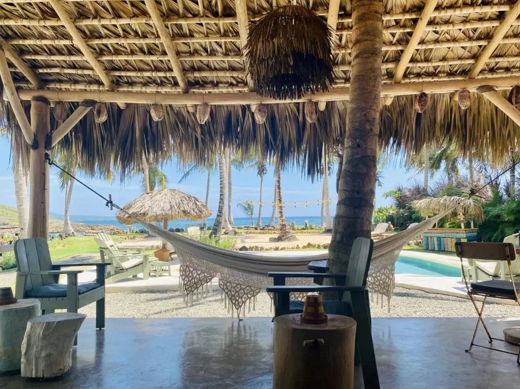 Open-air thatched hut with a hammock between palm trees, overlooking a beach and pool.