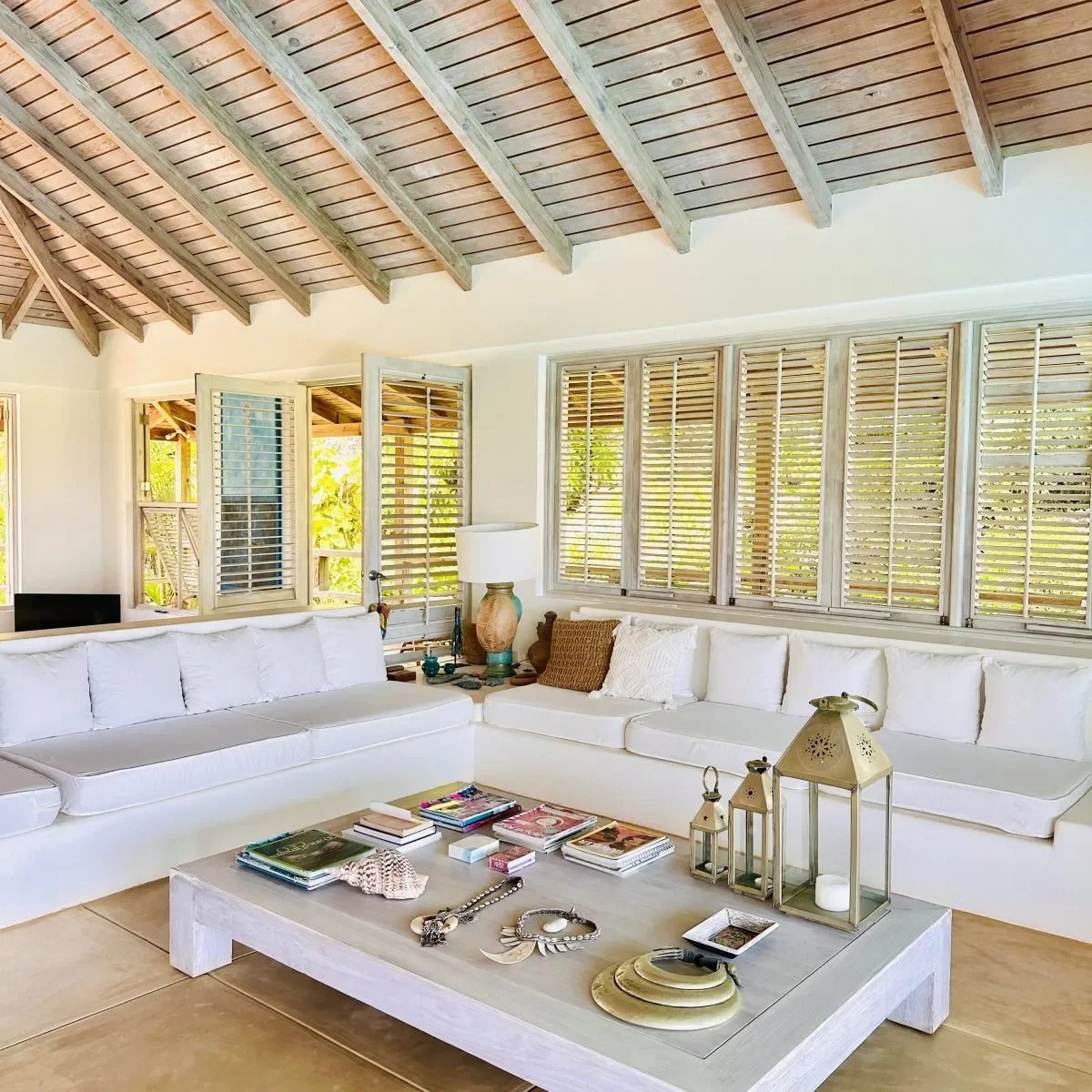 Bright coastal living room with a white sectional, light wood coffee table, and large window shutters, letting in daylight.