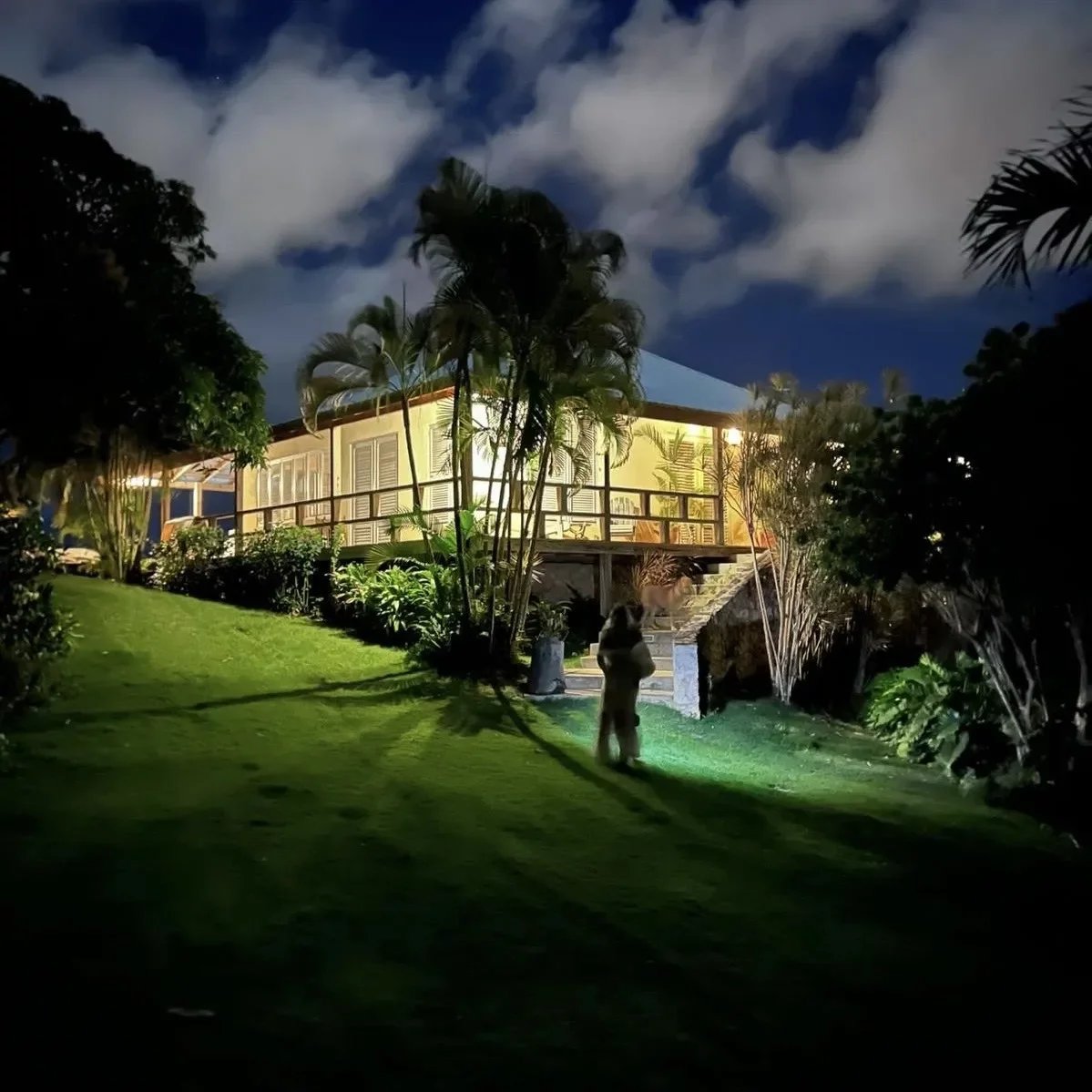 Night view of a yellow house with a wooden deck, palm trees, and a grassy lawn.