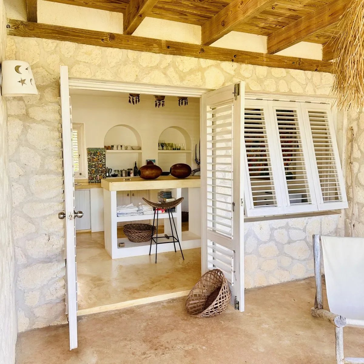 Open French doors reveal a sunny stone-walled outdoor kitchen with a white bar island, shelves, and wicker baskets on the floor and counter top sweat.