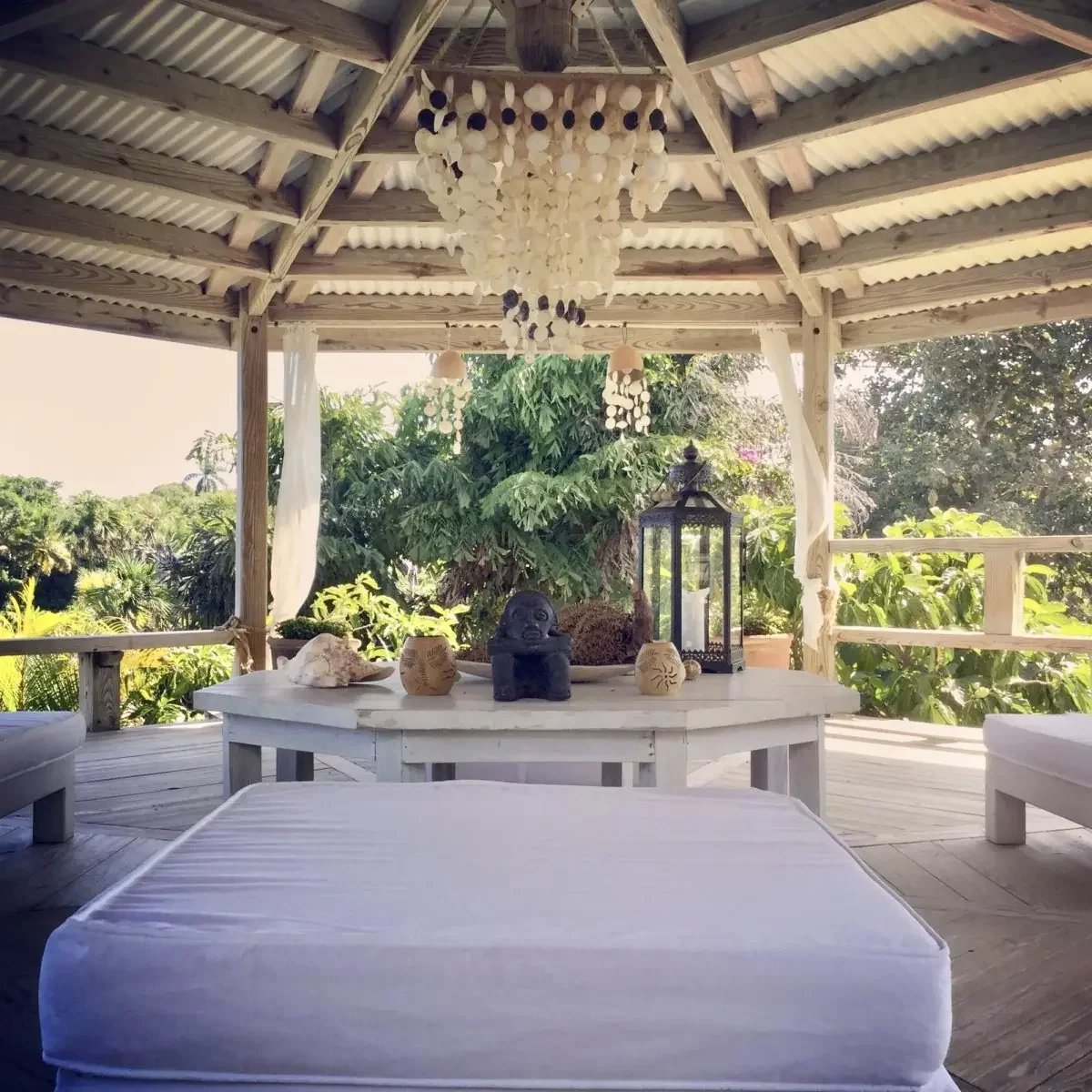 White wooden gazebo with a circular table and shell-inspired decor, lantern, and cushioned lounges in a tropical garden setting.