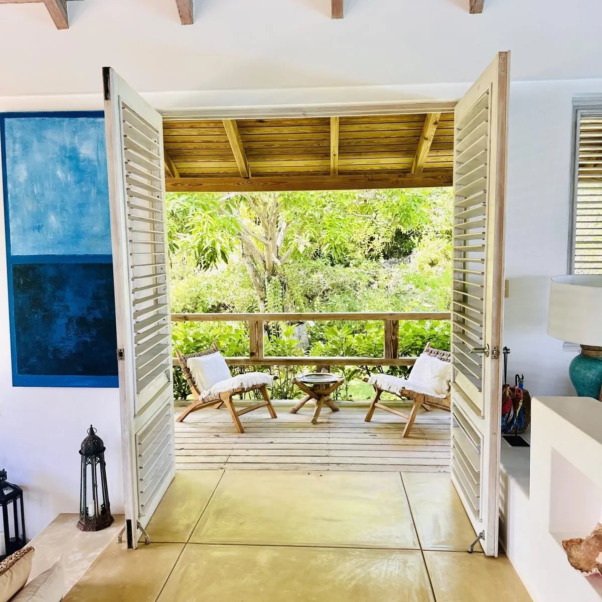 Open double doors reveal a wooden deck with two woven chairs and a small round table, overlooking a lush garden.