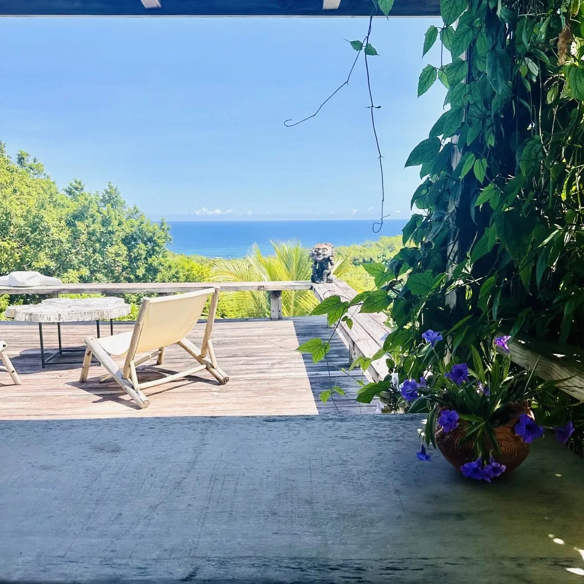 Ocean view from a wooden deck with two lounge chairs and a round table; a pot of purple flowers on the right and lush greenery surrounding the scene.