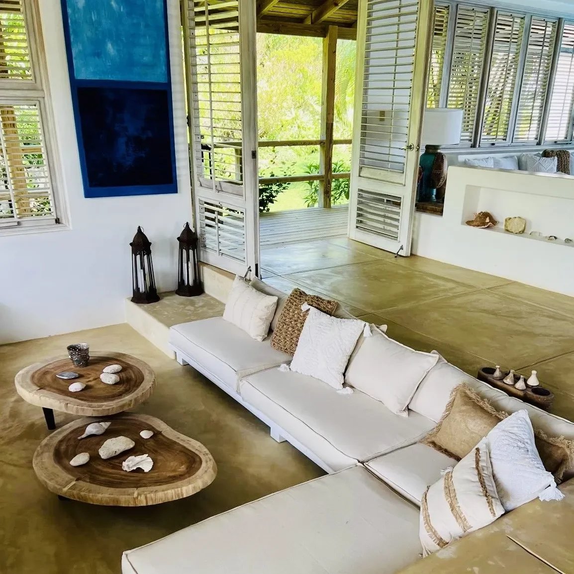 Bright living room with a white sectional sofa, beige cushions, two circular wooden coffee tables with shells, and open doors to a sunny outdoor area.