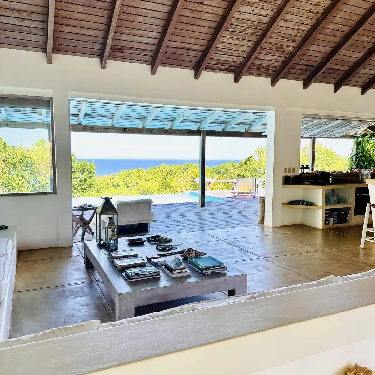 Open-plan living room with white sofas and a low coffee table, opening to a deck and the ocean view beyond.