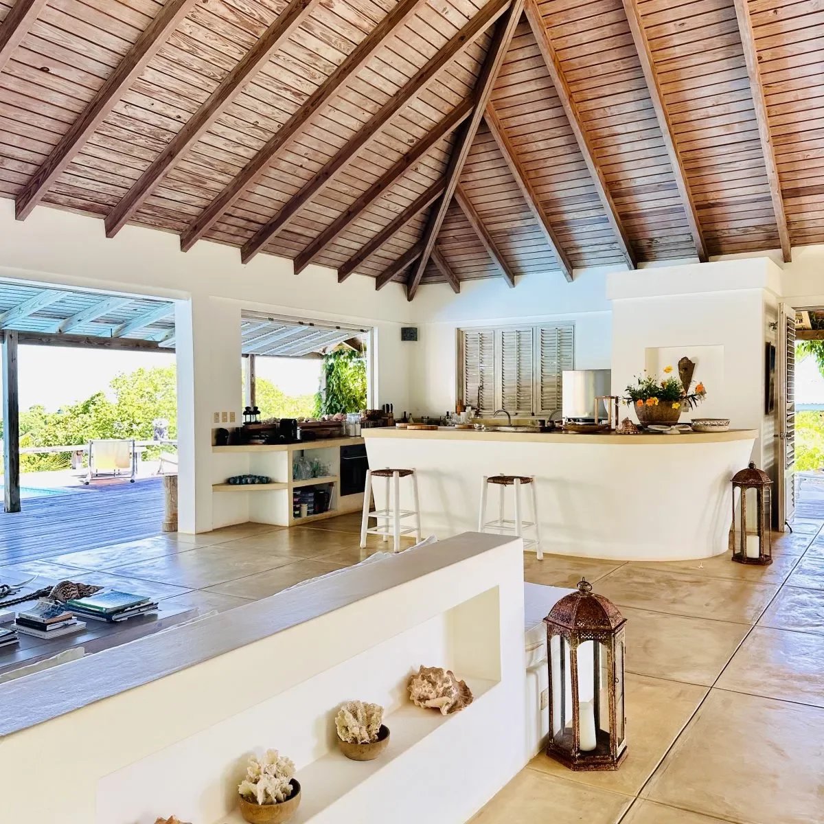 Open tropical kitchen with a curved white island, three stools, and a vaulted wooden ceiling overlooking a deck.