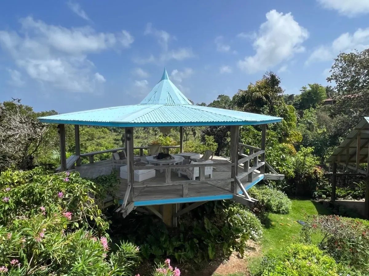 Open-air gazebo with a turquoise pyramid roof on a wooden platform, surrounded by lush tropical garden and seating. (Informative)