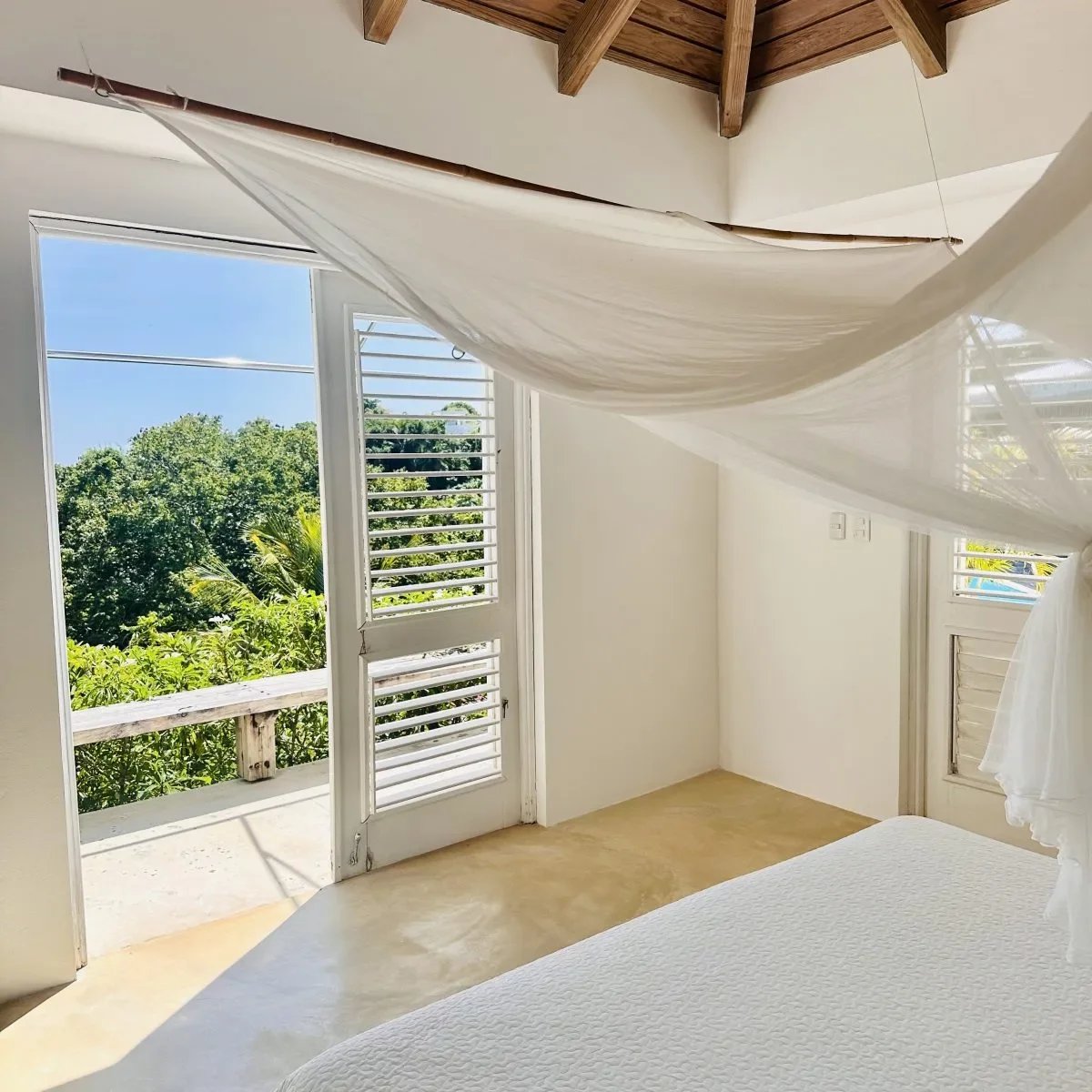 Sunlit bedroom with a white sheer canopy draped from wooden beams, open door to a balcony with green trees outside.