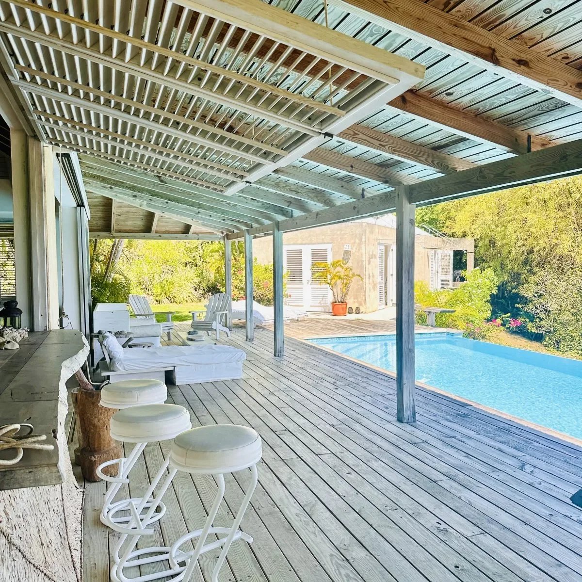 Covered backyard deck with white bar stools, lounge chairs, and a rectangular pool surrounded by greenery