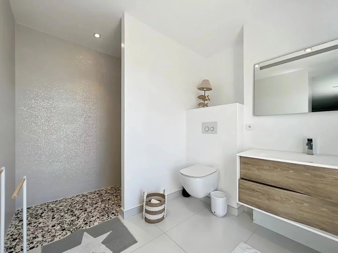 Minimalist bathroom with a wall-mounted toilet, white countertop vanity with wood drawers, and a walk-in shower with pebble floor. Inset decor includes a small basket and a star rug nearby.