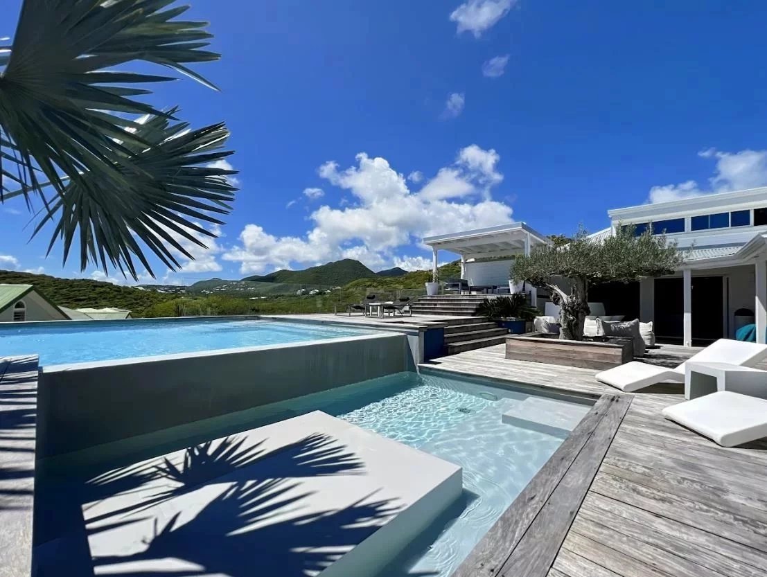 Luxury poolside terrace with a wooden deck, white lounge furniture, and bright blue sky over a modern villa.