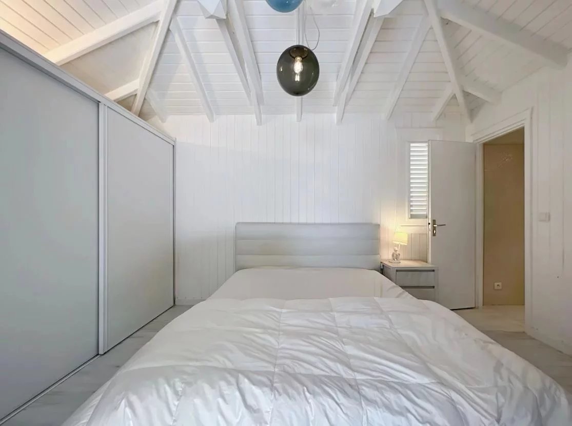 Minimalist white bedroom with a padded headboard bed, nightstand and lamp, and a sliding-door wardrobe along the left wall under a vaulted white ceiling.