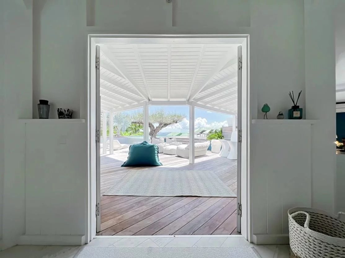 Open doors reveal a bright white patio with a teal cushion on a rug and white seating under a pergola, with a blue sky and ocean view in the background.
