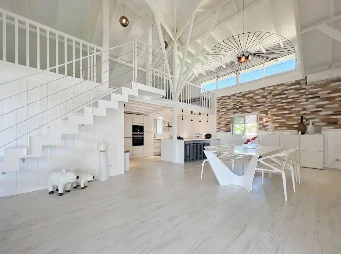 Bright open-plan white kitchen and dining area with a sculptural staircase and a large circular black wire pendant light above the table.