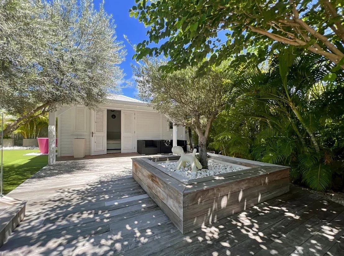 Sunlit backyard featuring a white shed with open doors, a wooden deck, and a raised wooden planter filled with white stones and an abstract sculpture under a tree.