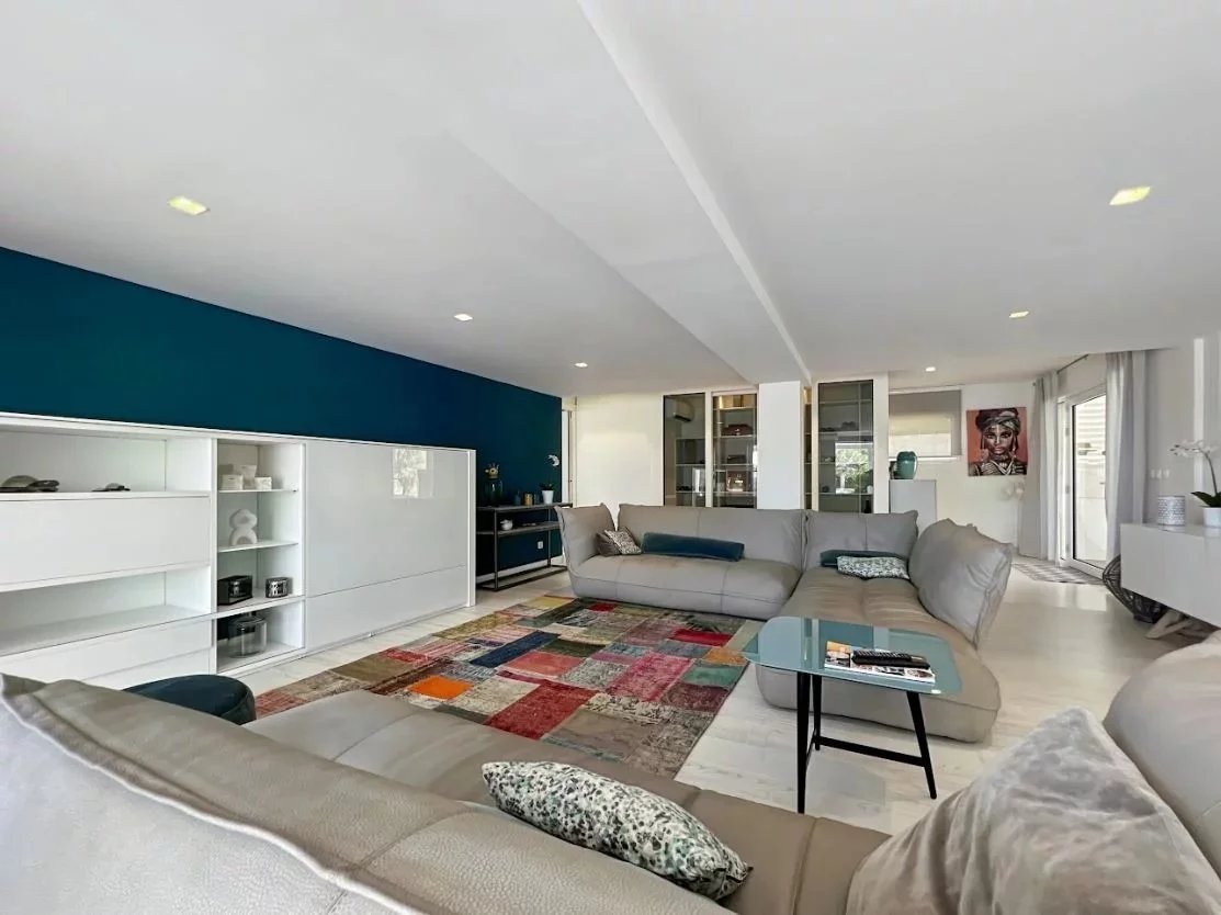 Modern open-plan living room with an L-shaped gray sectional, a colorful patchwork rug, and a teal accent wall along white built-in storage units; glass coffee table and decorative cushions add color.