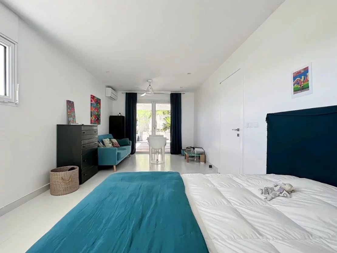 Bright bedroom with white walls, teal bedspread, and a seating area by the glass doors to a balcony.