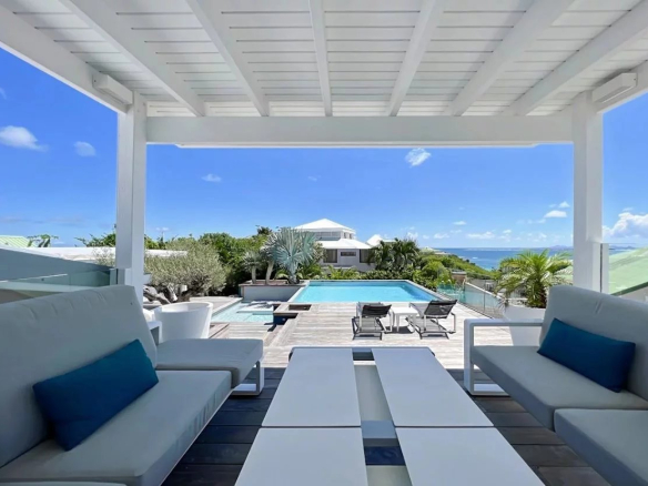 White sectional sofa with blue cushions under a shaded pergola overlooking a rectangular pool and the ocean beyond.