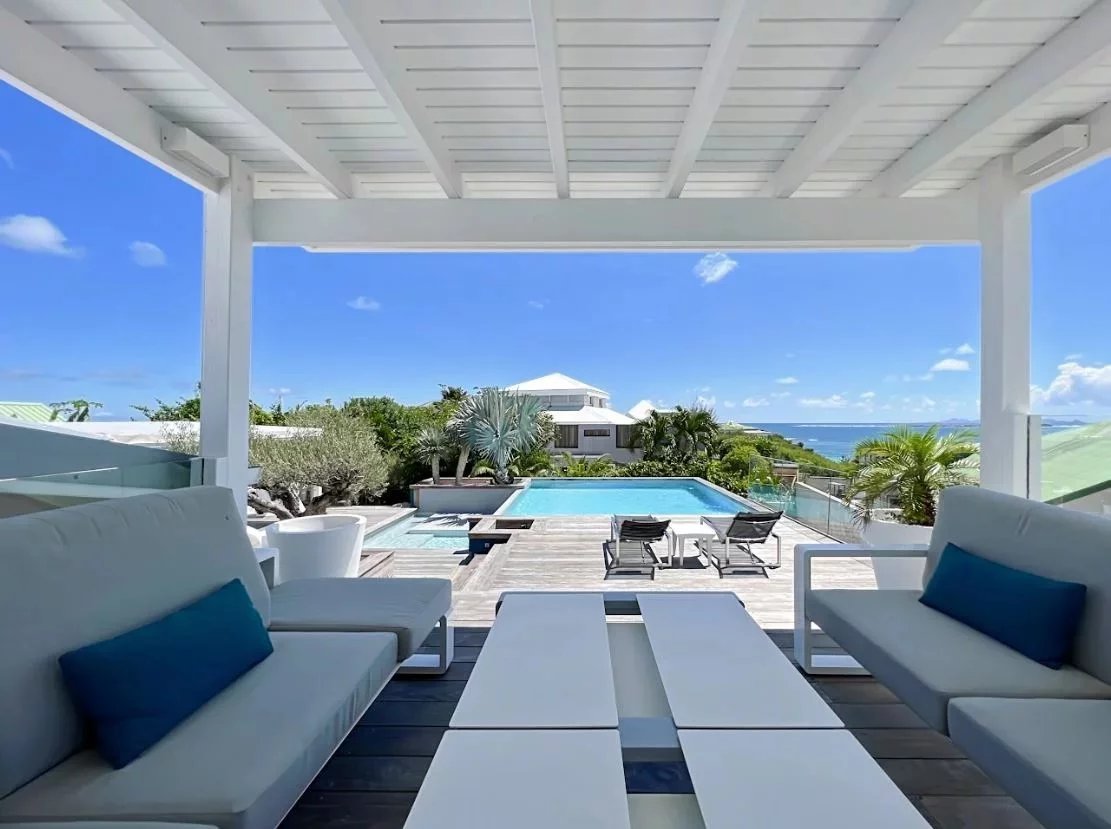 White sectional sofa with blue cushions under a shaded pergola overlooking a rectangular pool and the ocean beyond Propriétés clovis