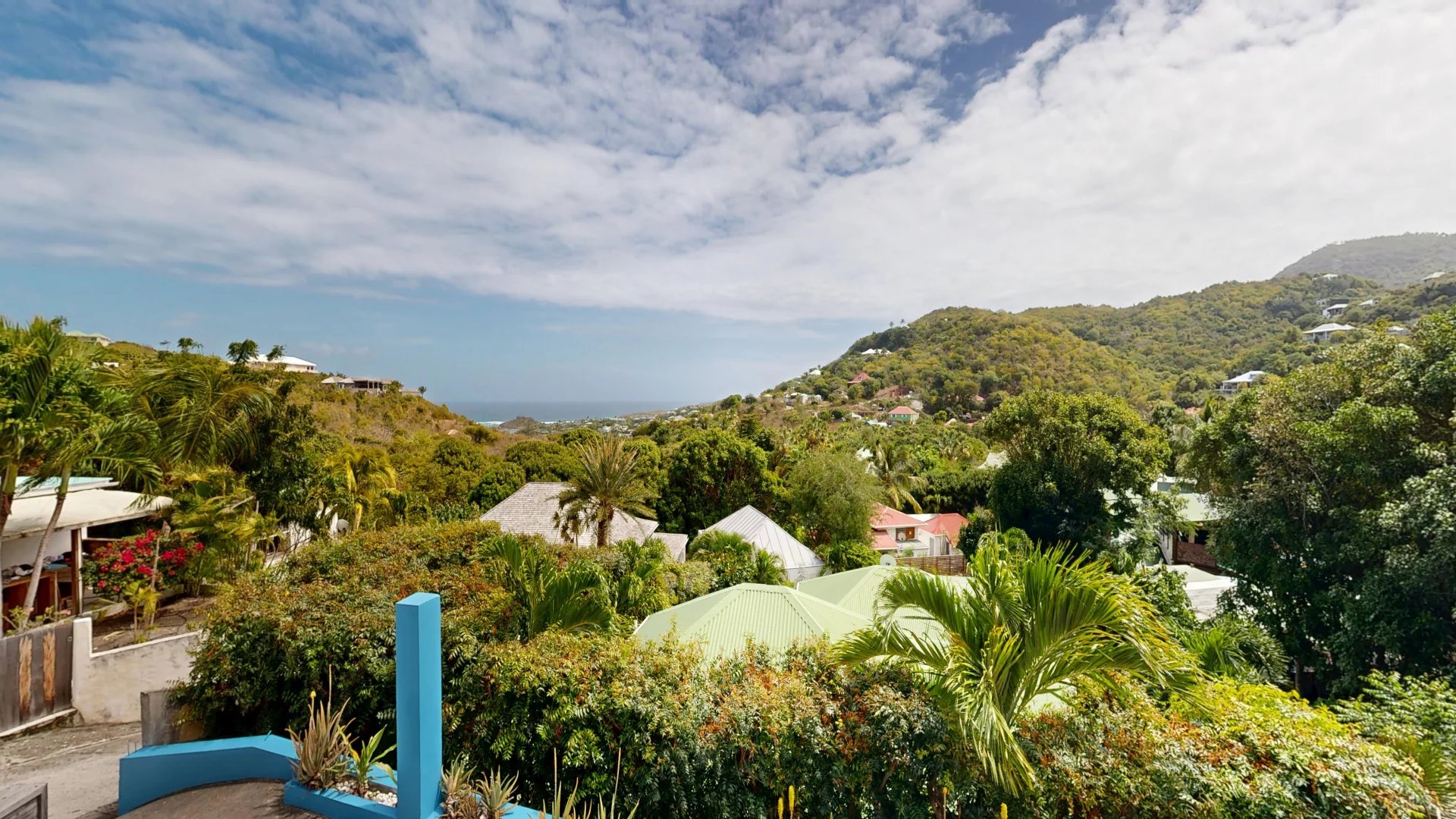 Tropical hillside village with palm trees, pastel rooftops, and the blue sea in the distance under a partly cloudy sky, lush greenery in the foreground.
