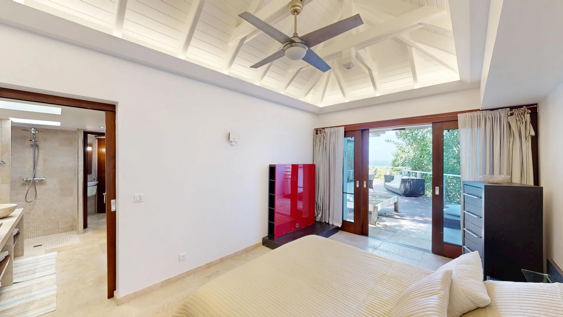 Bright bedroom with a white vaulted ceiling, ceiling fan, and glass doors opening to a balcony with outdoor seating area.