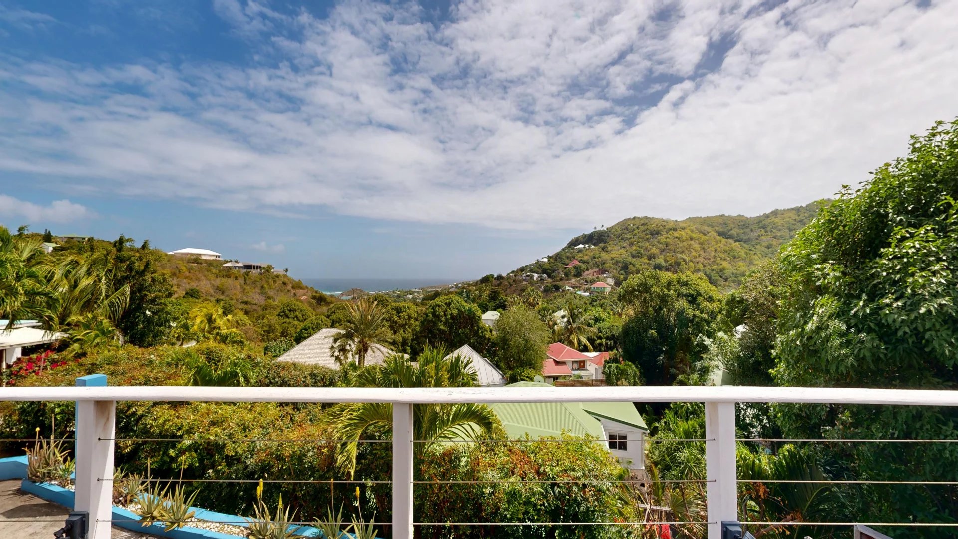 View from a balcony over tropical hills with green houses and a distant ocean under a blue sky with clouds