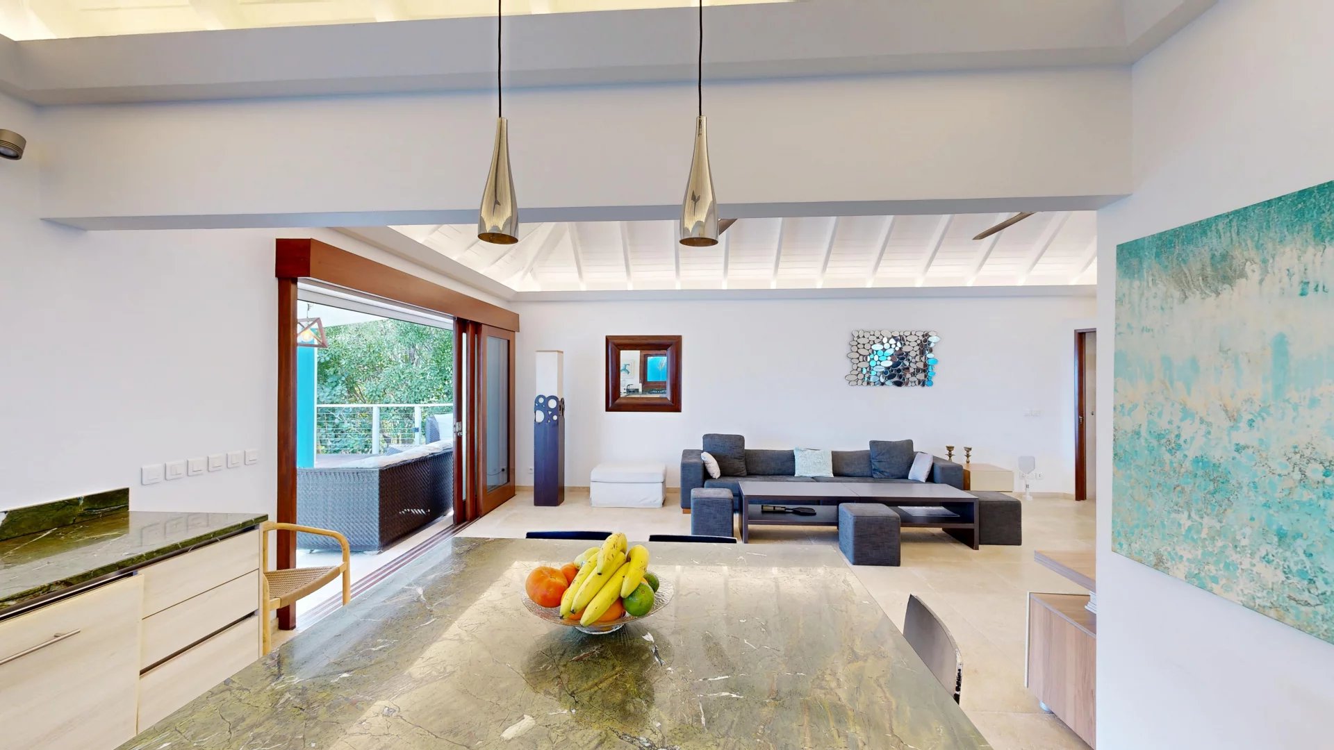 Open-plan living and kitchen space with a marble island, fruit bowl, and a gray sectional sofa facing a coffee table and wall art.